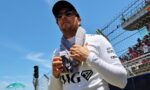 Man wearing sunglasses and a cap with a towel around his neck at a racetrack, near a chain-link fence and spectators.