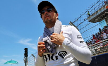 Man wearing sunglasses and a cap with a towel around his neck at a racetrack, near a chain-link fence and spectators.