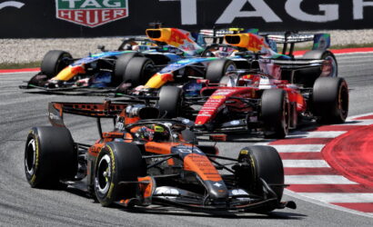A pack of Formula One cars races through a sharp corner, led by an orange McLaren with other colorful F1 cars close behind on the track.