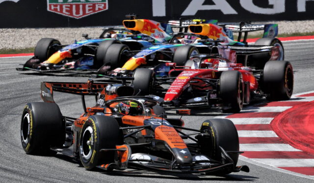 A pack of Formula One cars races through a sharp corner, led by an orange McLaren with other colorful F1 cars close behind on the track.