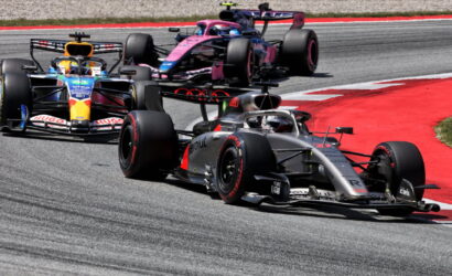 Three Formula 1 cars race through a tight corner on a gray race track with red runoff edging the bend; the lead car is silver and black.