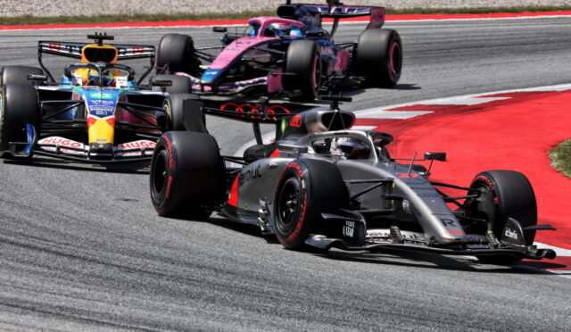 Three Formula 1 cars race through a tight corner on a gray race track with red runoff edging the bend; the lead car is silver and black.