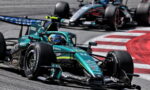 Teal Formula 1 car leading the pack on a track, driver in helmet, close behind another race car around a red-and-white curbing edge.