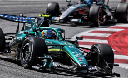 Teal Formula 1 car leading the pack on a track, driver in helmet, close behind another race car around a red-and-white curbing edge.