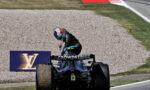 Racing driver in black suit standing on back of a Formula 1 car on a gravel runoff at a track, rear wing labeled 'CROWDSTRIKE' and a Louis Vuitton sign in the background.