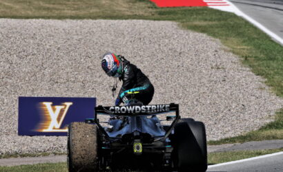 Racing driver in black suit standing on back of a Formula 1 car on a gravel runoff at a track, rear wing labeled 'CROWDSTRIKE' and a Louis Vuitton sign in the background.