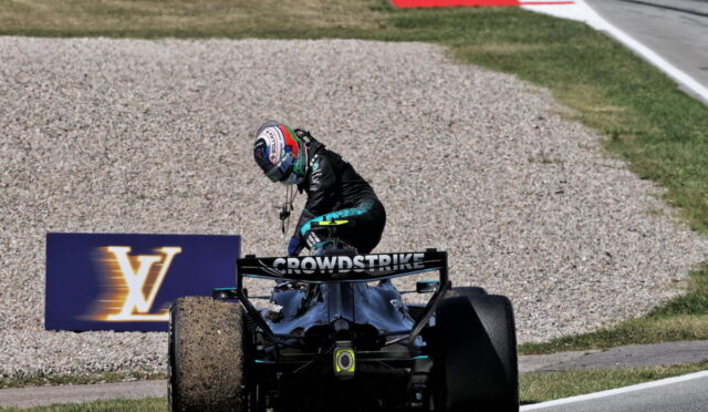 Racing driver in black suit standing on back of a Formula 1 car on a gravel runoff at a track, rear wing labeled 'CROWDSTRIKE' and a Louis Vuitton sign in the background.