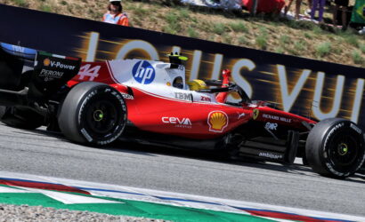 Red Ferrari Formula 1 car speeding along a track with sponsor logos (Shell, HP) and a Louis Vuitton banner in the background, spectators nearby.