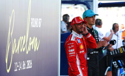 Two Formula 1 drivers in team suits stand by a pit wall, the red Ferrari driver talking on a phone while the blue-capped Mercedes driver watches nearby.