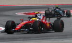 Red Ferrari Formula 1 car leads a tight midfield pack around a sweeping race corner on the track, sponsor logos visible on bodywork