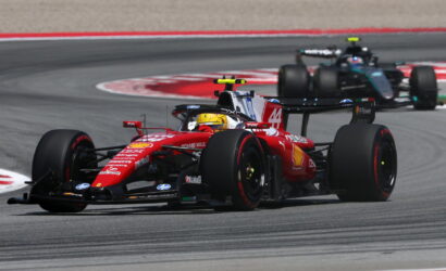 Red Ferrari Formula 1 car leads a tight midfield pack around a sweeping race corner on the track, sponsor logos visible on bodywork