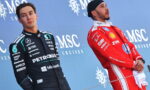 Two Formula 1 drivers on a podium against a blue backdrop, one in a black Petronas racing suit, the other in a red race suit.