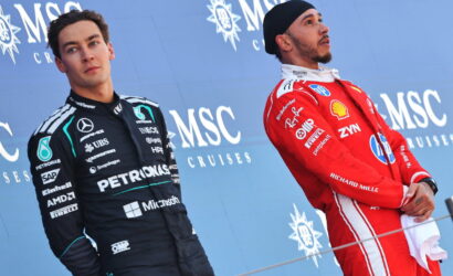 Two Formula 1 drivers on a podium against a blue backdrop, one in a black Petronas racing suit, the other in a red race suit.