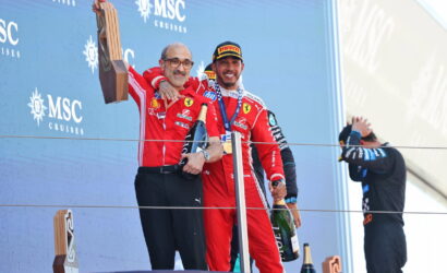 Two Ferrari drivers celebrate on the podium, one raises a wooden trophy while the other holds a champagne bottle and wears a medal, against a blue MSC Cruises backdrop.