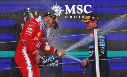 Race car driver in red suit sprays champagne from a Moët bottle on the podium as another driver in dark gear watches, MSC Cruises backdrop behind them.