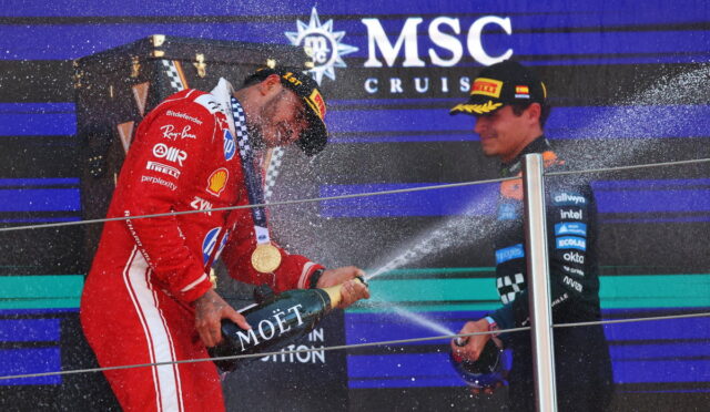Race car driver in red suit sprays champagne from a Moët bottle on the podium as another driver in dark gear watches, MSC Cruises backdrop behind them.