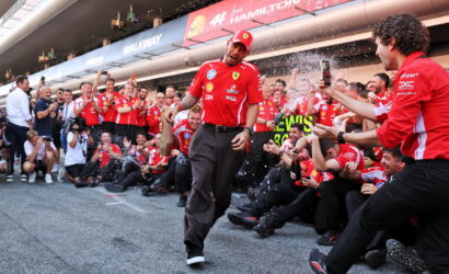 Red-clad Ferrari team celebrations: a man walks forward as teammates spray champagne in a pit lane.