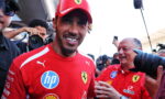 Ferrari driver smiling in red team shirt, surrounded by photographers at a media event.