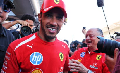 Ferrari driver smiling in red team shirt, surrounded by photographers at a media event.