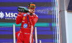 Racecar driver in a red suit on a podium spraying champagne from a Moët bottle.