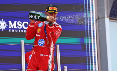 Racecar driver in a red suit on a podium spraying champagne from a Moët bottle.