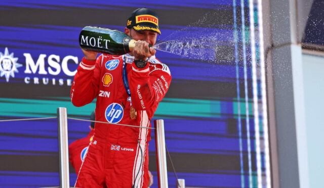 Racecar driver in a red suit on a podium spraying champagne from a Moët bottle.