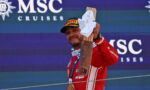 Race car driver in a red suit raises a wooden trophy and towel on a podium, wearing a gold medal, with an MSC Cruises backdrop.