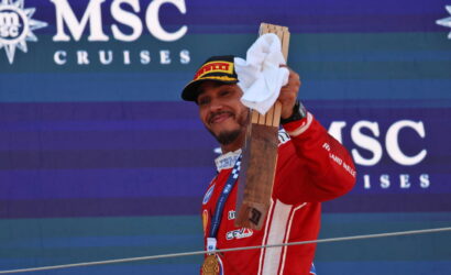 Race car driver in a red suit raises a wooden trophy and towel on a podium, wearing a gold medal, with an MSC Cruises backdrop.