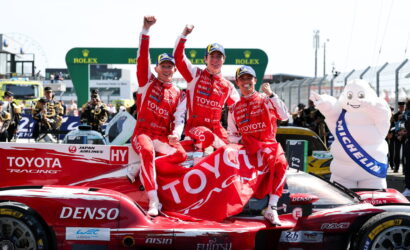 Three Toyota Racing drivers celebrate on a red race car, raising their fists with a large Toyota banner in the background.]"