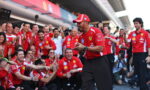 Ferrari team at a victory celebration in the pit lane, crowd cheering around a smiling man in a red Ferrari shirt and cap clapping.