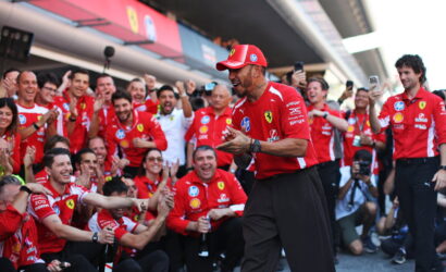 Ferrari team at a victory celebration in the pit lane, crowd cheering around a smiling man in a red Ferrari shirt and cap clapping.