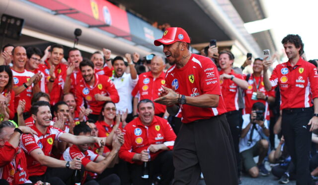 Ferrari team at a victory celebration in the pit lane, crowd cheering around a smiling man in a red Ferrari shirt and cap clapping.