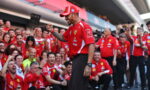 Ferrari team member in red shirt and cap, raising a fist while surrounded by cheering teammates and fans at a race pit area.