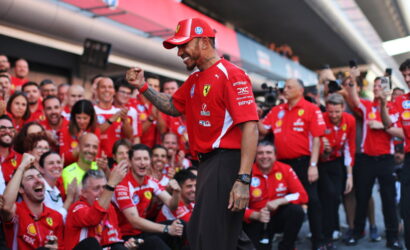 Ferrari team member in red shirt and cap, raising a fist while surrounded by cheering teammates and fans at a race pit area.