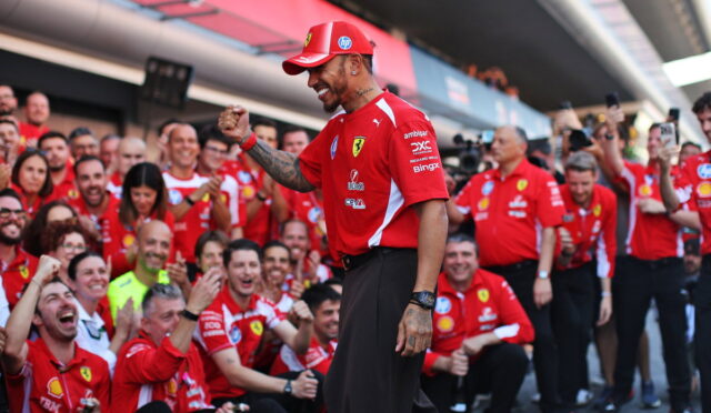 Ferrari team member in red shirt and cap, raising a fist while surrounded by cheering teammates and fans at a race pit area.