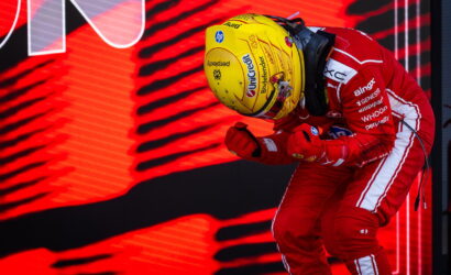 F1 driver in a red racing suit and yellow helmet leans forward in a victory celebration against a red LED backdrop simple, bold colors.