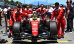 Ferrari Formula 1 car being pushed by red-suited crew in the pit lane with engineers and photographers nearby, motorsport banners in the background.
