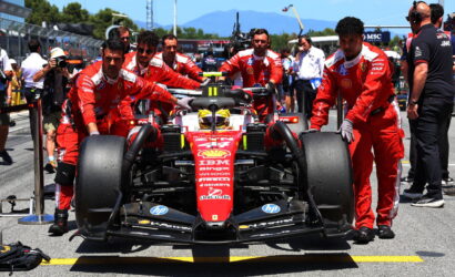 Ferrari Formula 1 car being pushed by red-suited crew in the pit lane with engineers and photographers nearby, motorsport banners in the background.