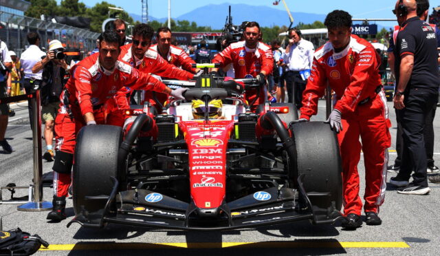 Ferrari Formula 1 car being pushed by red-suited crew in the pit lane with engineers and photographers nearby, motorsport banners in the background.