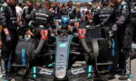 Mercedes F1 car in the pit with crew surrounding the cockpit, driver in helmet preparing for a pit stop or launch, team logos visible (Petronas, Mercedes) and front wing close to the ground.
