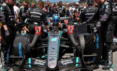Mercedes F1 car in the pit with crew surrounding the cockpit, driver in helmet preparing for a pit stop or launch, team logos visible (Petronas, Mercedes) and front wing close to the ground.