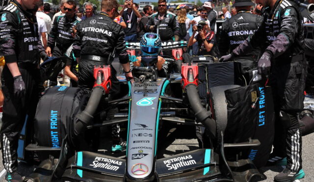 Mercedes F1 car in the pit with crew surrounding the cockpit, driver in helmet preparing for a pit stop or launch, team logos visible (Petronas, Mercedes) and front wing close to the ground.