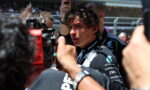 Young race car driver in a dark Petronas racing suit surrounded by reporters and cameras at a pit lane press moment