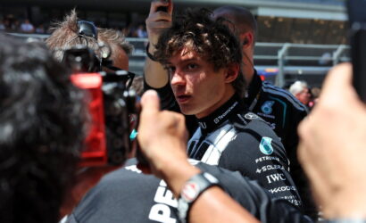 Young race car driver in a dark Petronas racing suit surrounded by reporters and cameras at a pit lane press moment
