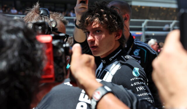 Young race car driver in a dark Petronas racing suit surrounded by reporters and cameras at a pit lane press moment