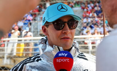 Male interviewer wearing a metallic silver jacket and blue cap with a Mercedes logo speaks into a Sky Sport microphone, stadium crowd in the background.