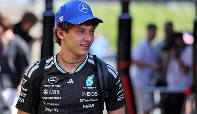 Young man in a black Mercedes racing jersey and a blue cap with the Mercedes star, standing outdoors at a event.