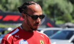 Close-up of a man with braided hair wearing a red Ferrari shirt and dark sunglasses, looking to the side outdoors.