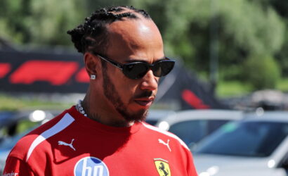 Close-up of a man with braided hair wearing a red Ferrari shirt and dark sunglasses, looking to the side outdoors.