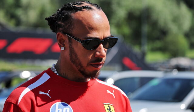 Close-up of a man with braided hair wearing a red Ferrari shirt and dark sunglasses, looking to the side outdoors.